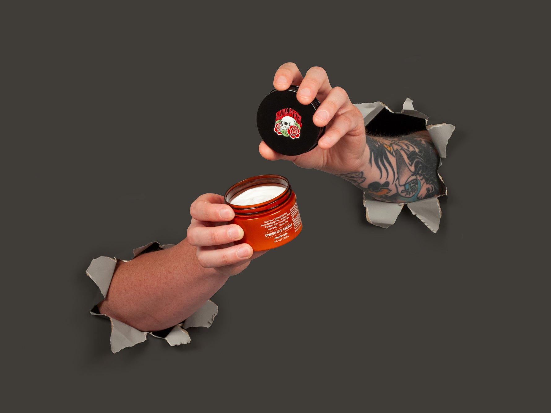 Two hands breaking through gray paper wall holding the under eye cream product