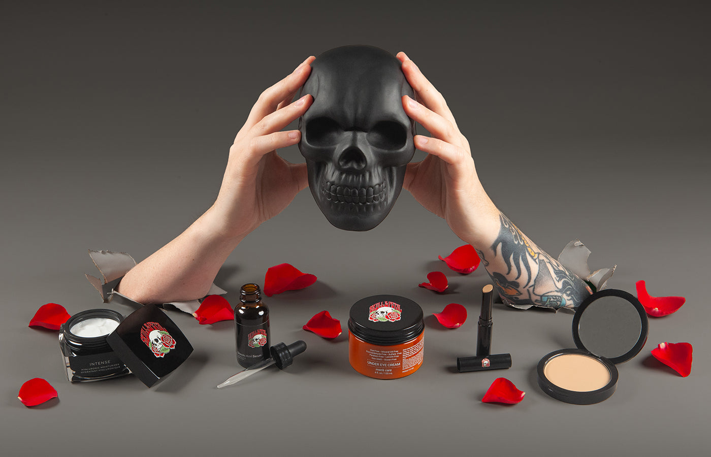 Skull & Petal products along with rose petals on a gray surface with two hands breaking through the surface, holding a black skull